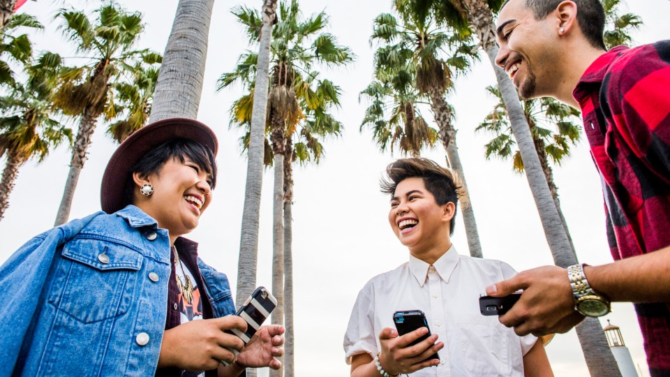 A group of three people together laughing with phones in their hands, surrounded by palm trees.