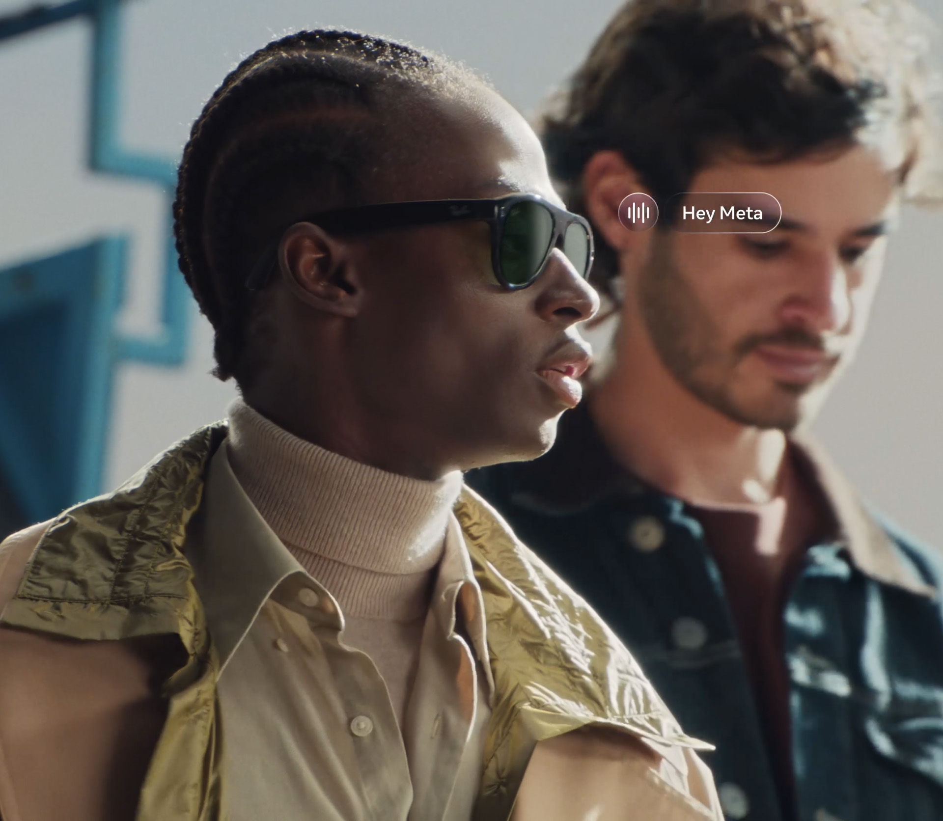 A man wearing dark Ray-Ban Meta glasses with green lenses, with an on-screen Hey Meta voice prompt beside him.